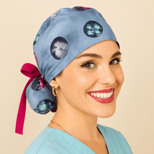 "Galaxy scrub cap with planet and space print on blue fabric, ponytail style tied with a pink ribbon bow. Modeled by a smiling woman in a light blue scrub top with gold hoop earrings and red lipstick. Everyday Heroes medical cap, reusable and washable cotton surgical hat for women nurses, doctors, surgeons, and dental staff. Comfortable satin lined scrub cap designed for healthcare professionals."