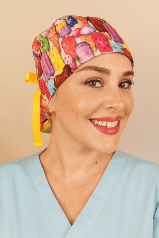 Ice creams scrub ponytail hat for women featuring colorful ice cream and popsicle print on cotton fabric, tied with a yellow ribbon bow and Everyday Heroes label. Satin lined scrub cap with buttons for mask comfort, modeled by a smiling nurse in light blue scrubs. Personalized surgical hat custom with name, reusable and washable medical cap. Perfect gift for nurses, doctors, and healthcare professionals."
