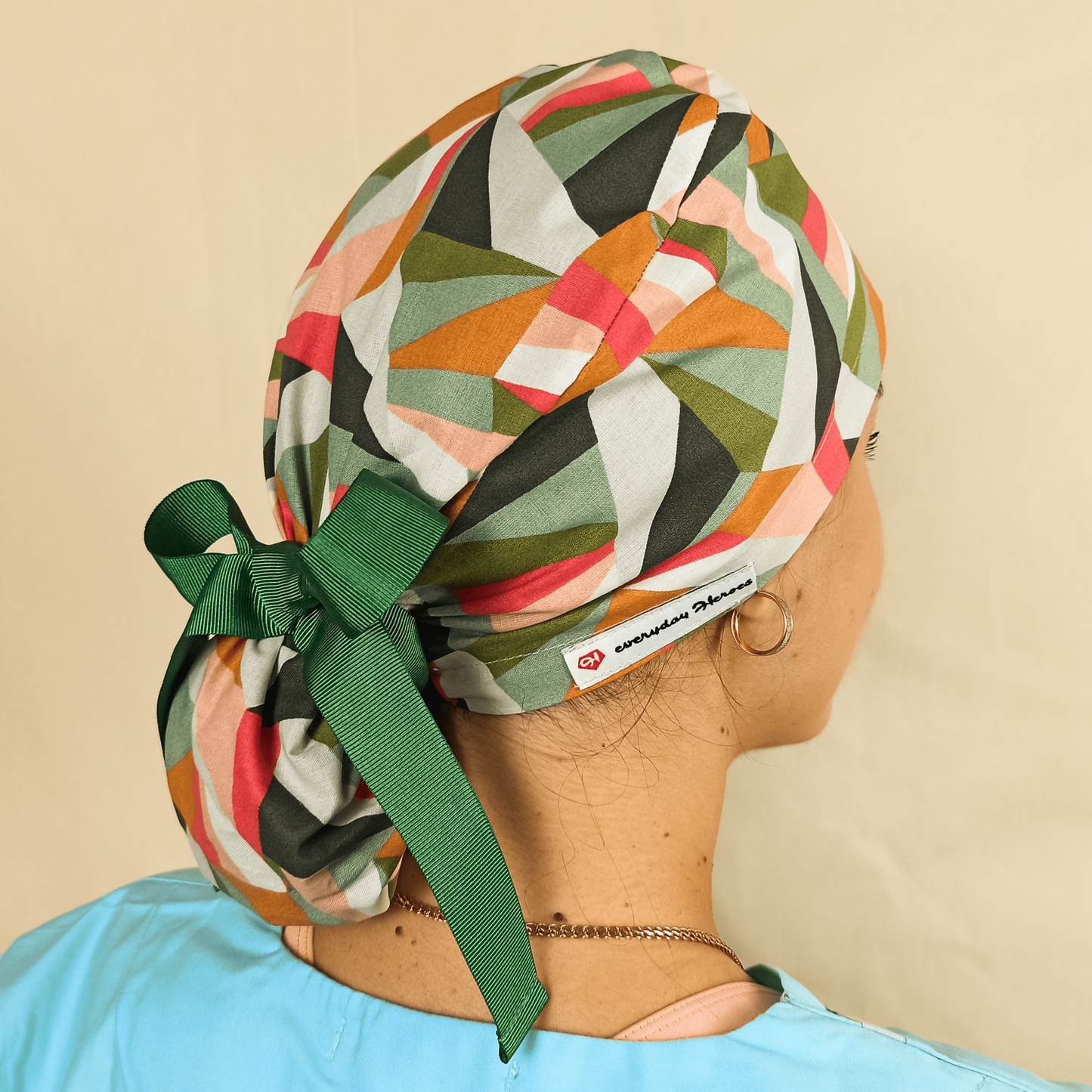 Nurse scrub cap with colorful geometrical abstract print in green and coral, satin lined ponytail scrub cap with buttons for mask comfort. Modeled by a smiling woman in a light blue scrub top with hoop earrings, featuring a tie back style for secure fit. Custom surgical cap for women, reusable and washable, ideal for nurses, doctors, surgeons, and healthcare staff. Personalized medical hat, perfect nurse gift.