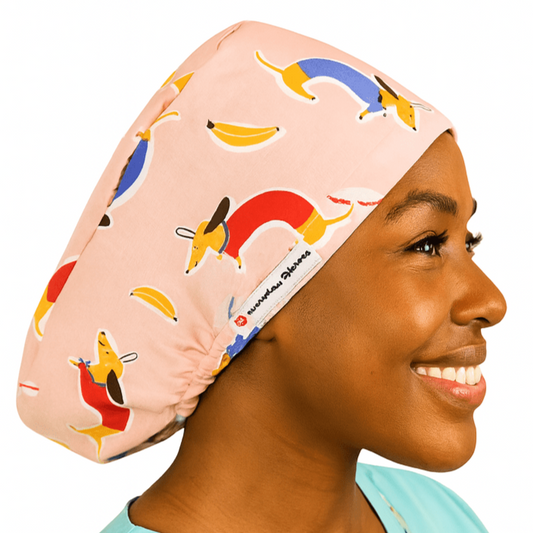 Smiling woman wearing a pink dachshund vet scrub cap with colorful sausage dog and banana pattern. Custom satin-lined euro scrub hat for veterinarians, surgeons, and nurses. Playful weenie dog print adds personality and comfort for animal care professionals.