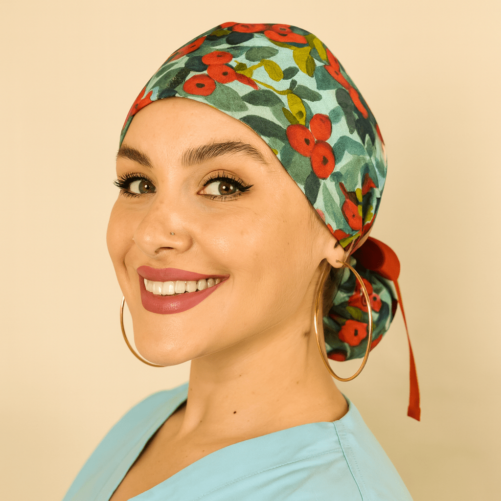 Floral scrub cap with red and green watercolor flower print, satin lined ponytail style, tied with a red ribbon bow and Everyday Heroes label. Modeled by a smiling woman in a light blue scrub top with gold hoop earrings and natural makeup. Comfortable reusable surgical hat for nurses, doctors, surgeons, and dental staff. Washable cotton medical cap designed for women healthcare professionals.