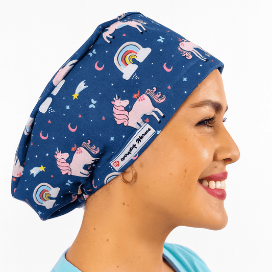 Smiling woman wearing a navy euro scrub cap with pink unicorns, rainbows, and stars print. Fun pediatric cartoon scrub hat, customizable with name embroidery. Ideal for nurses, surgeons, and pediatric staff working in the operating room.