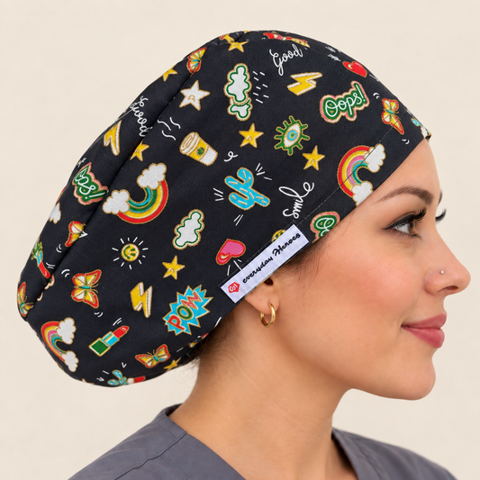 A woman in profile wearing a black scrub cap with a colorful cartoon-style print of rainbows, stars, lightning bolts, and medical-themed doodles. 