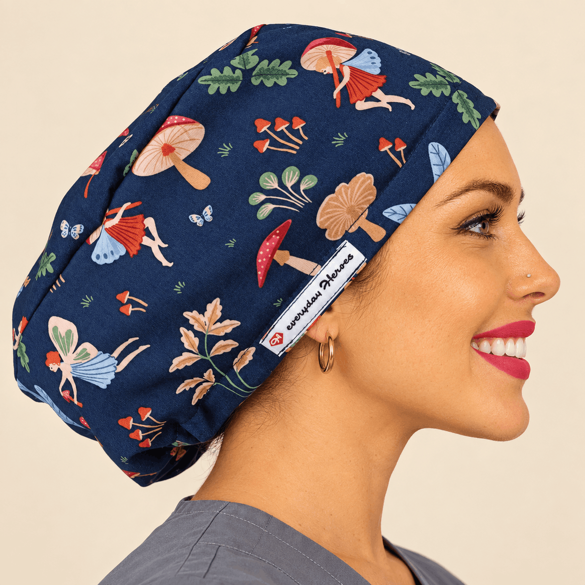 A woman in profile wearing a navy blue scrub cap with a whimsical mushroom and fairy print. The fabric features red, beige, green, and orange illustrations of mushrooms, leaves, and fairies.