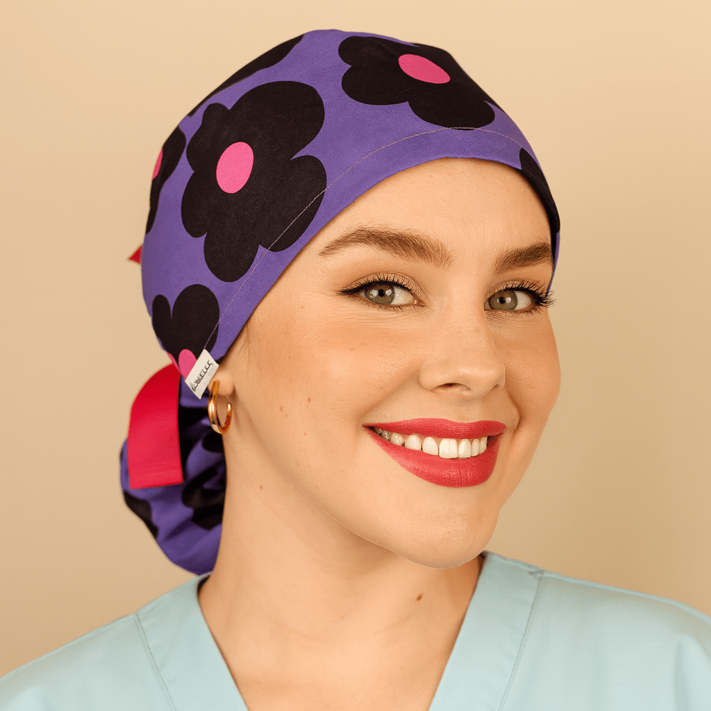 Purple scrub cap with bold black floral print and pink centers, ponytail tie back style with bright ribbon and Everyday Heroes label. Modeled by a smiling woman in a light blue scrub top with gold hoop earrings and natural makeup. Washable cotton surgical hat for nurses, doctors, surgeons, and dental staff. Comfortable satin lined scrub cap designed for women healthcare professionals.