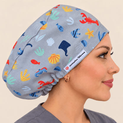 A woman in profile wearing a light gray scrub cap with a colorful underwater-themed print of crabs, fish, shells, and sea plants. A white brand tag reading 'surgical headwear' is visible on the side of the cap.