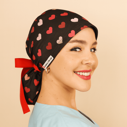 Black scrub cap with red and pink heart print, satin lined, featuring a red bow tie back and white Everyday Heroes label. Modeled by a smiling nurse wearing a light blue scrub top, this surgical hat is designed for women doctors, nurses, and surgeons. Comfortable medical cap with ponytail holder, reusable and washable, ideal for hospital, clinic, or dental staff.