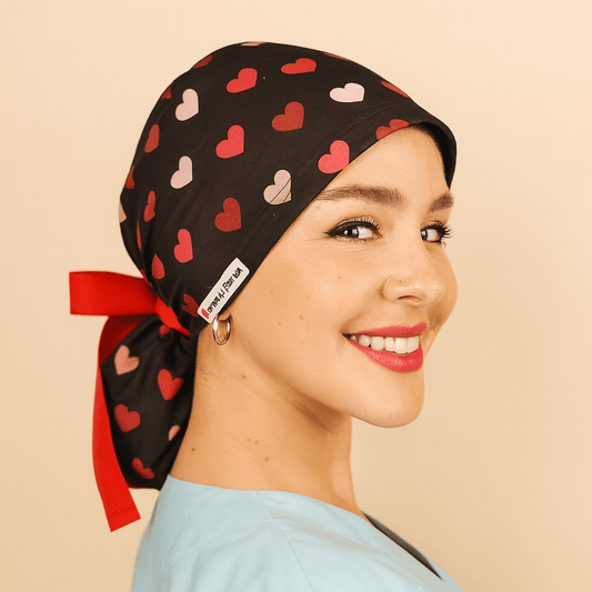 Black scrub cap with red and pink heart print, satin lined, featuring a red bow tie back and white Everyday Heroes label. Modeled by a smiling nurse wearing a light blue scrub top, this surgical hat is designed for women doctors, nurses, and surgeons. Comfortable medical cap with ponytail holder, reusable and washable, ideal for hospital, clinic, or dental staff.