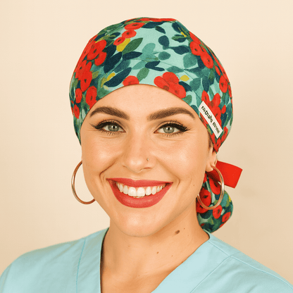 Floral scrub cap with red and green watercolor flower print, satin lined ponytail style, tied with a red ribbon bow and Everyday Heroes label. Modeled by a smiling woman in a light blue scrub top with gold hoop earrings and natural makeup. Comfortable reusable surgical hat for nurses, doctors, surgeons, and dental staff. Washable cotton medical cap designed for women healthcare professionals.
