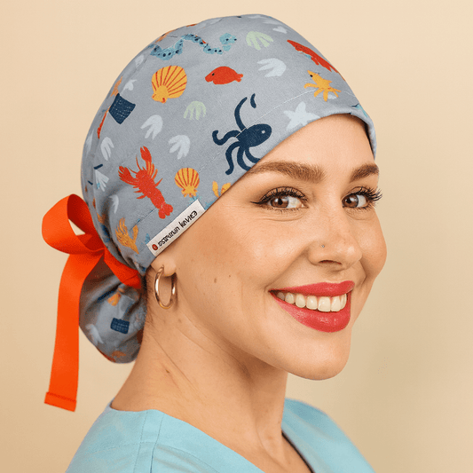 Gray scrub cap with colorful ocean sea life print featuring octopus, fish, shells, and coral. Ponytail tie back style with bright orange ribbon bow and Everyday Heroes label. Modeled by a smiling woman in a light blue scrub top with gold hoop earrings and bold red lipstick. Washable cotton surgical hat for nurses, doctors, and surgeons. Comfortable reusable medical scrub cap designed for women healthcare staff."