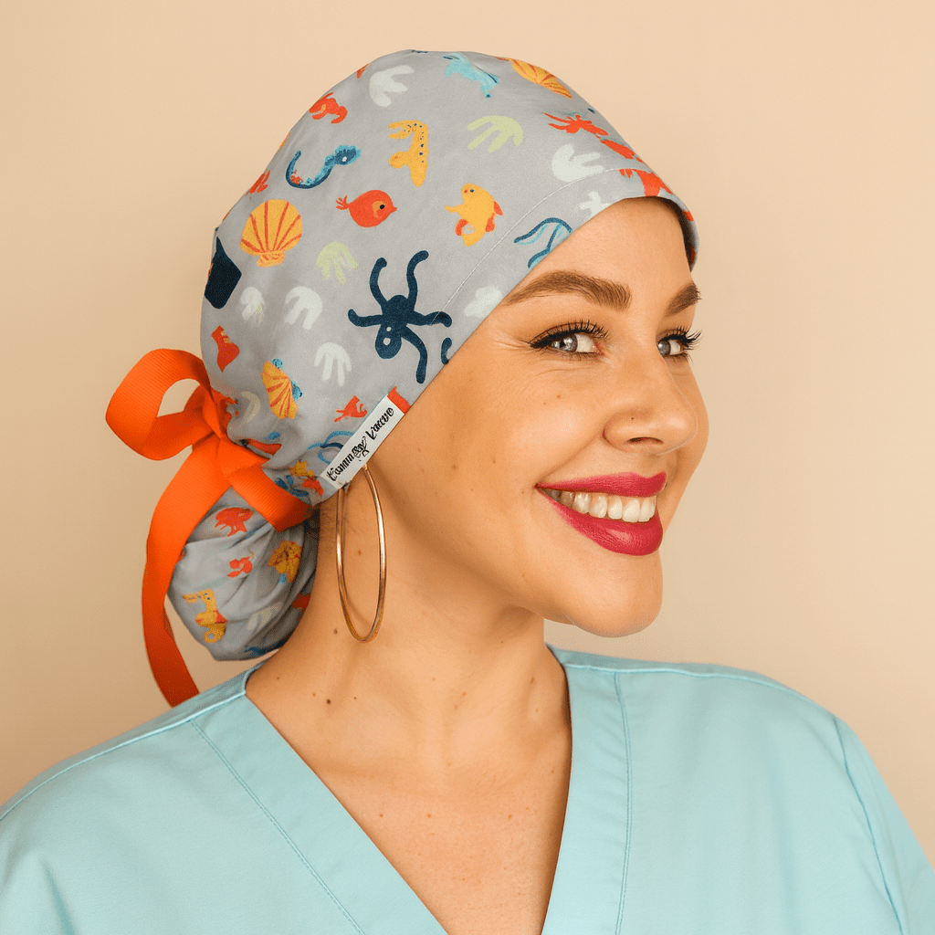Gray scrub cap with colorful ocean sea life print featuring octopus, fish, shells, and coral. Ponytail tie back style with bright orange ribbon bow and Everyday Heroes label. Modeled by a smiling woman in a light blue scrub top with gold hoop earrings and bold red lipstick. Washable cotton surgical hat for nurses, doctors, and surgeons. Comfortable reusable medical scrub cap designed for women healthcare staff."