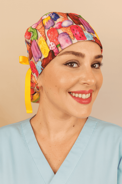 Ice creams scrub ponytail hat for women featuring colorful ice cream and popsicle print on cotton fabric, tied with a yellow ribbon bow and Everyday Heroes label. Satin lined scrub cap with buttons for mask comfort, modeled by a smiling nurse in light blue scrubs. Personalized surgical hat custom with name, reusable and washable medical cap. Perfect gift for nurses, doctors, and healthcare professionals."
