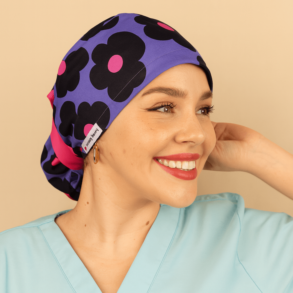 Purple scrub cap with bold black floral print and pink centers, ponytail tie back style with bright ribbon and Everyday Heroes label. Modeled by a smiling woman in a light blue scrub top with gold hoop earrings and natural makeup. Washable cotton surgical hat for nurses, doctors, surgeons, and dental staff. Comfortable satin lined scrub cap designed for women healthcare professionals.