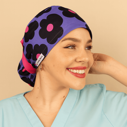 Purple scrub cap with bold black floral print and pink centers, ponytail tie back style with bright ribbon and Everyday Heroes label. Modeled by a smiling woman in a light blue scrub top with gold hoop earrings and natural makeup. Washable cotton surgical hat for nurses, doctors, surgeons, and dental staff. Comfortable satin lined scrub cap designed for women healthcare professionals.