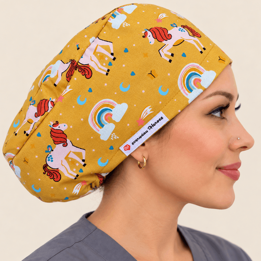 A woman in profile wearing a mustard yellow scrub cap with a whimsical print of white unicorns, red manes, pastel rainbows, and small stars. 