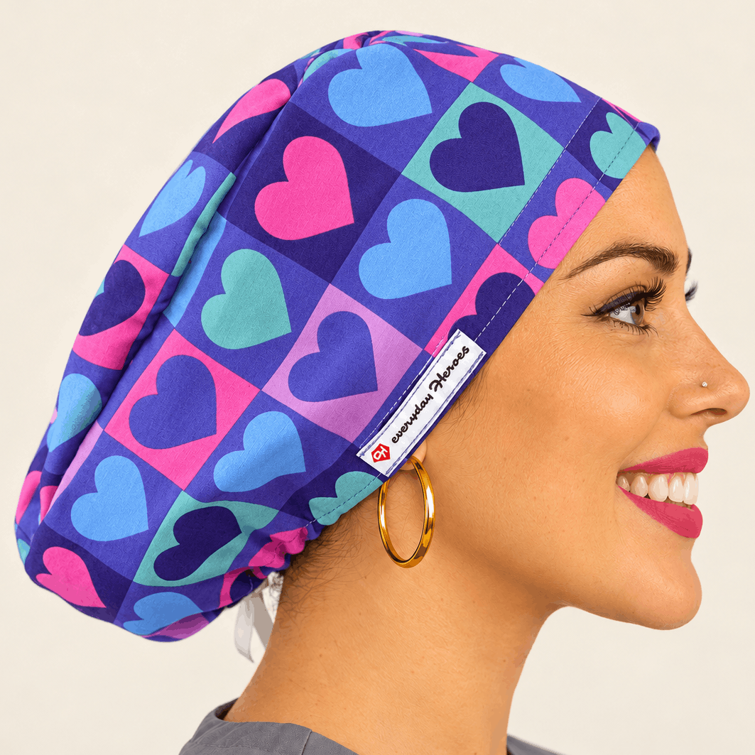 Beautiful brunette healthcare worker wearing purple and pink hearts pattern bouffant scrub cap with Everyday Heroes label, smiling in three-quarter view against white background. Premium medical scrub hat with colorful heart print design, adjustable fit, professional nursing cap for women. High-quality surgical cap with satin lining, handmade in Greece, perfect for nurses, doctors, and medical professionals.