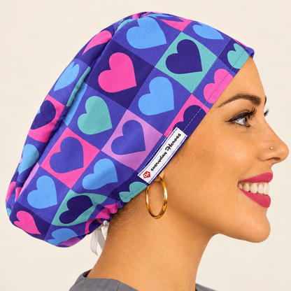 Beautiful brunette healthcare worker wearing purple and pink hearts pattern bouffant scrub cap with Everyday Heroes label, smiling in three-quarter view against white background. Premium medical scrub hat with colorful heart print design, adjustable fit, professional nursing cap for women. High-quality surgical cap with satin lining, handmade in Greece, perfect for nurses, doctors, and medical professionals.