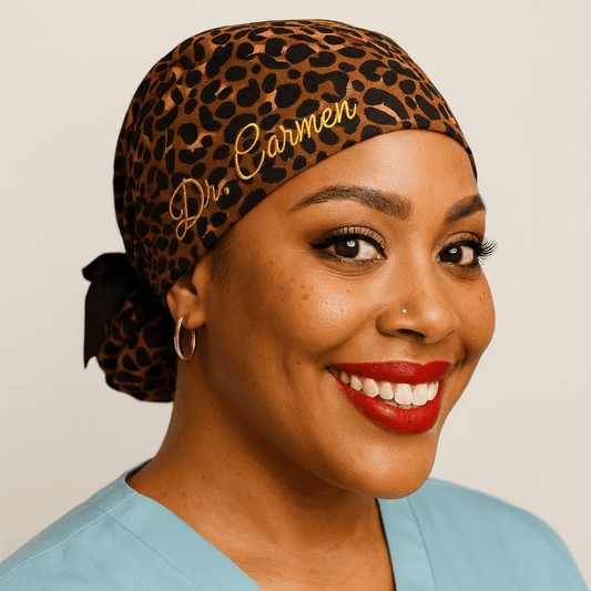 Black female healthcare professional wearing leopard print ponytail scrub cap with "Dr. Carmen" gold embroidery, black ribbon bow, smiling in light blue scrubs. Premium satin-lined surgical cap with custom embroidered name in metallic gold thread. Adjustable tie-back design for nurses, doctors, surgical technologists. Cheetah leopard animal print pattern on beige background. Gift for nurse, gift idea for doctor, personalized medical headwear, professional healthcare scrub cap.