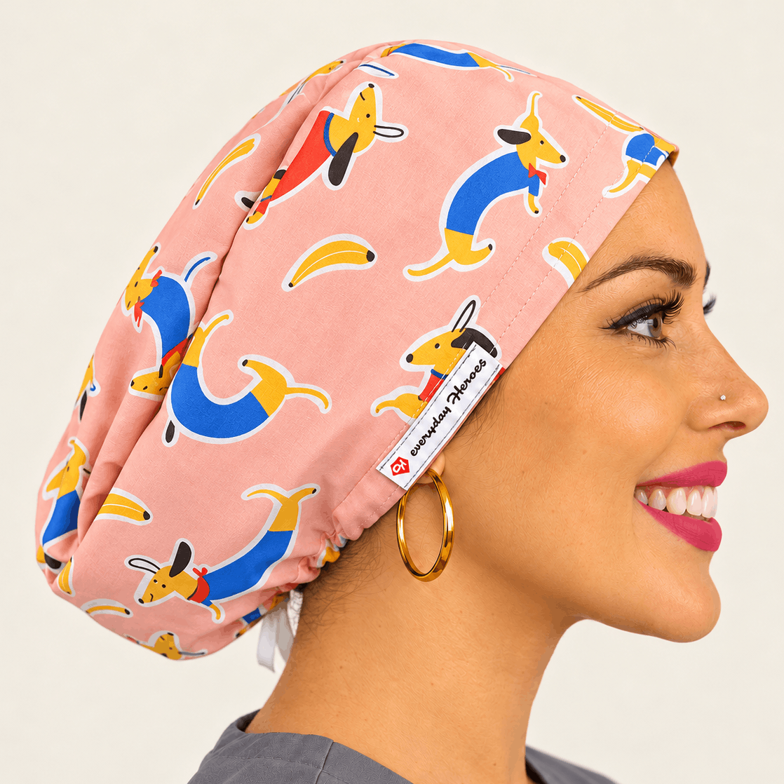 A smiling woman in profile wearing a pink scrub cap with a colorful dachshund print. The scrub cap features a white tag with the brand name