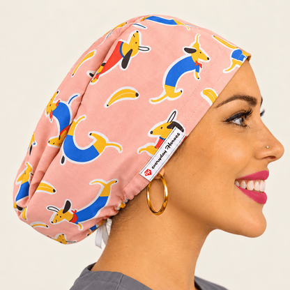 A smiling woman in profile wearing a pink scrub cap with a colorful dachshund print. The scrub cap features a white tag with the brand name