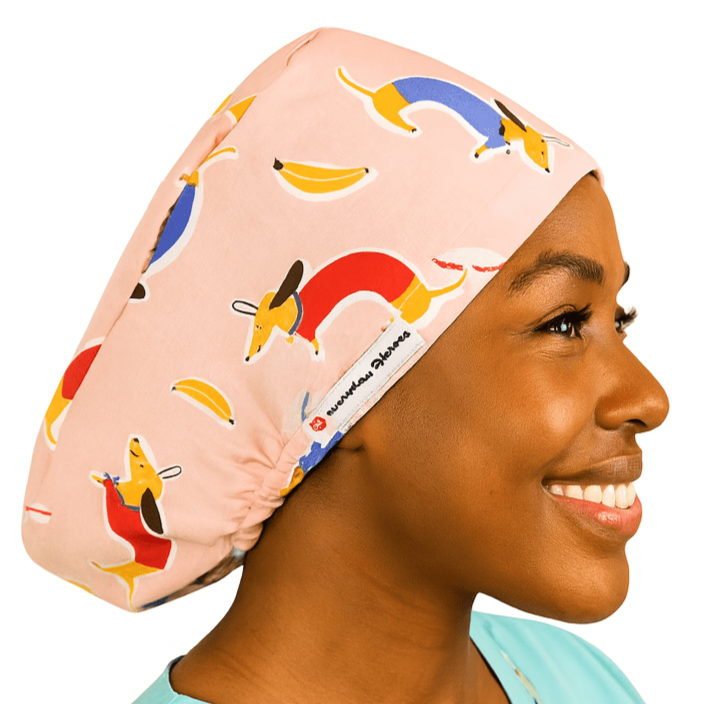 Smiling woman wearing a pink dachshund vet scrub cap with colorful sausage dog and banana pattern. Custom satin-lined euro scrub hat for veterinarians, surgeons, and nurses. Playful weenie dog print adds personality and comfort for animal care professionals.