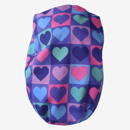 Beautiful brunette healthcare worker wearing purple and pink hearts pattern bouffant scrub cap with Everyday Heroes label, smiling in three-quarter view against white background. Premium medical scrub hat with colorful heart print design, adjustable fit, professional nursing cap for women. High-quality surgical cap with satin lining, handmade in Greece, perfect for nurses, doctors, and medical professionals.