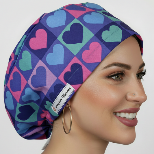 Beautiful brunette healthcare worker wearing purple and pink hearts pattern bouffant scrub cap with Everyday Heroes label, smiling in three-quarter view against white background. Premium medical scrub hat with colorful heart print design, adjustable fit, professional nursing cap for women. High-quality surgical cap with satin lining, handmade in Greece, perfect for nurses, doctors, and medical professionals.