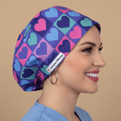 Beautiful brunette healthcare worker wearing purple and pink hearts pattern bouffant scrub cap with Everyday Heroes label, smiling in three-quarter view against white background. Premium medical scrub hat with colorful heart print design, adjustable fit, professional nursing cap for women. High-quality surgical cap with satin lining, handmade in Greece, perfect for nurses, doctors, and medical professionals.
