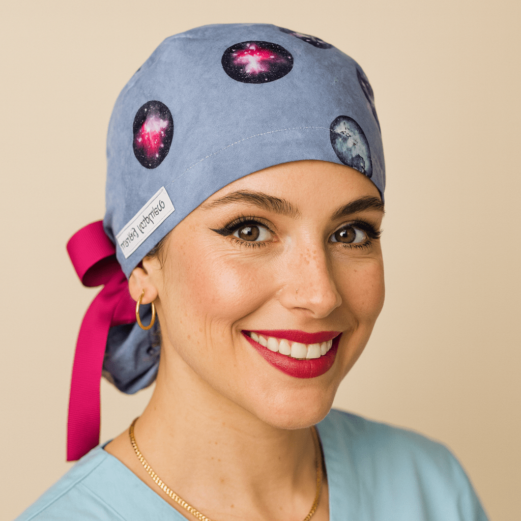 "Galaxy scrub cap with planet and space print on blue fabric, ponytail style tied with a pink ribbon bow. Modeled by a smiling woman in a light blue scrub top with gold hoop earrings and red lipstick. Everyday Heroes medical cap, reusable and washable cotton surgical hat for women nurses, doctors, surgeons, and dental staff. Comfortable satin lined scrub cap designed for healthcare professionals."