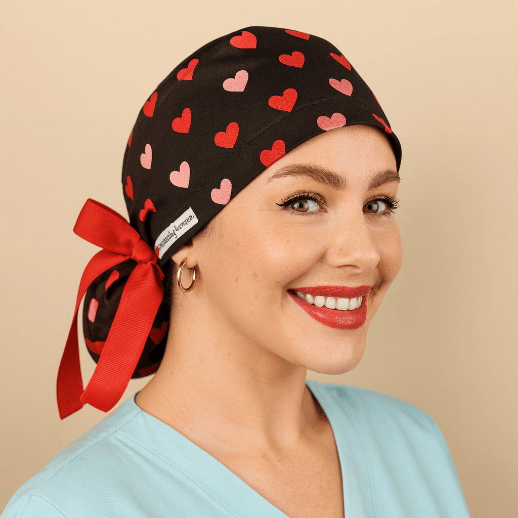 Black scrub cap with red and pink heart print, satin lined, featuring a red bow tie back and white Everyday Heroes label. Modeled by a smiling nurse wearing a light blue scrub top, this surgical hat is designed for women doctors, nurses, and surgeons. Comfortable medical cap with ponytail holder, reusable and washable, ideal for hospital, clinic, or dental staff.