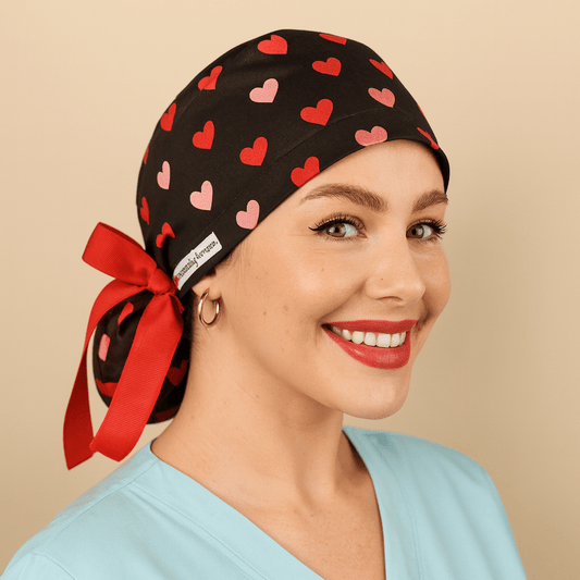 Black scrub cap with red and pink heart print, satin lined, featuring a red bow tie back and white Everyday Heroes label. Modeled by a smiling nurse wearing a light blue scrub top, this surgical hat is designed for women doctors, nurses, and surgeons. Comfortable medical cap with ponytail holder, reusable and washable, ideal for hospital, clinic, or dental staff.