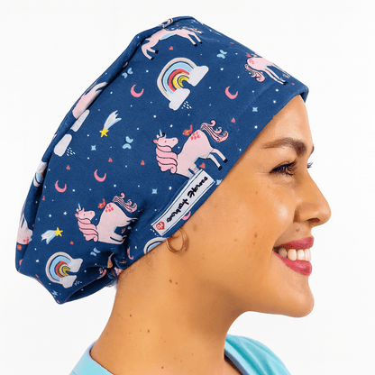 Smiling woman wearing a navy euro scrub cap with pink unicorns, rainbows, and stars print. Fun pediatric cartoon scrub hat, customizable with name embroidery. Ideal for nurses, surgeons, and pediatric staff working in the operating room.